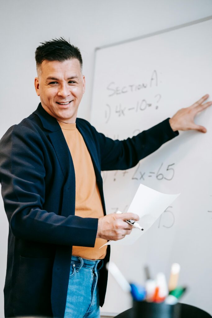 Friendly teacher explains equations on a whiteboard in a classroom setting.